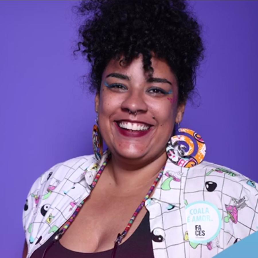 Foto da professora Cecí Romera, uma mulher negra usando um coque afro, maquiagem, brincos grandes e um colar, uma camisa branca quadriculada com vários pequenos desenhos e uma blusinha preta por dentro.
