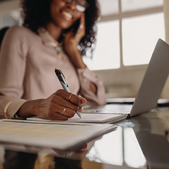 Foto de uma mulher negra de óculos sentada em uma mesa na frente de um notebook e escrevendo em um caderno.
