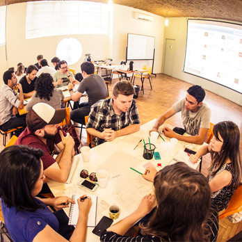 Sala de aula com várias mesas e cerca de 6 estudantes sentados em cada uma fazendo curso de UX Design, com folhas, celulares, post-its e outros materiais.