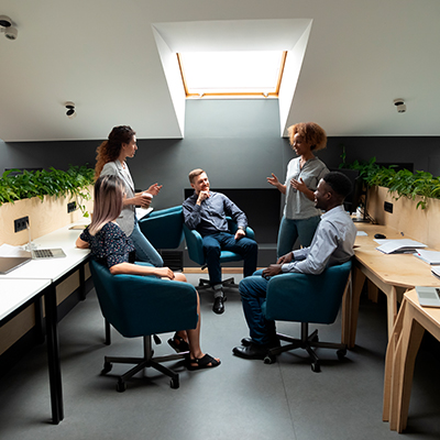 Foto de um escritório com bancadas com plantas e computadores na esquerda e na direita, e no centro cinco pessoas discutindo sobre trabalho.
