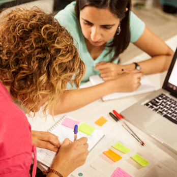 Mesa com duas estudantes sentadas fazendo curso de UX Design, com folhas, notebook, post-its e outros materiais.