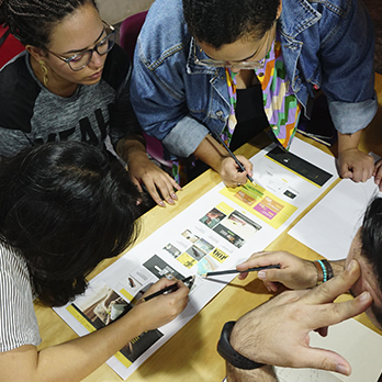 Uma mesa de madeira com quatro pessoas ao redor fazendo uma atividade do curso de Arquitetura de Informação, analisando uma interface que está impressa em um longo papel.