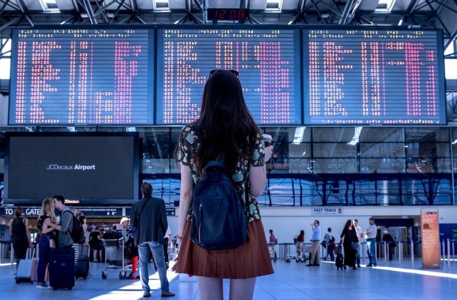 Foto de uma mulher de costas, usando mochila e saia, olhando para o painel de saídas e chegadas de vôos em um aeroporto, representando o curso Carreira de UX na Gringa.