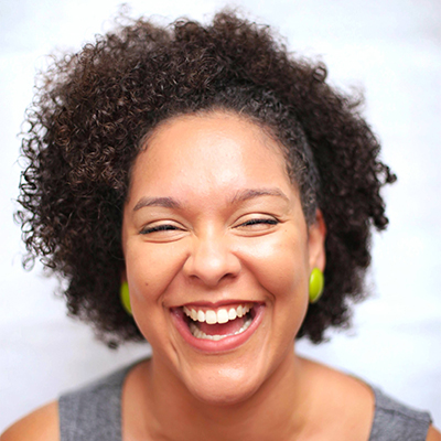 Foto de Aline Santos, apresentadora da DEXCONF 2024. Uma mulher negra de pele clara com cabelo crespo estilo black power, usando brincos verdes e sorrindo com os olhos levemente fechados.