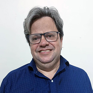 Foto de Guilherme Santa Rosa, palestrante da DEXCONF 2024. Um homem branco de cabelos grisalhos curtos usando óculos preto e camisa azul escuro, e sorrindo.