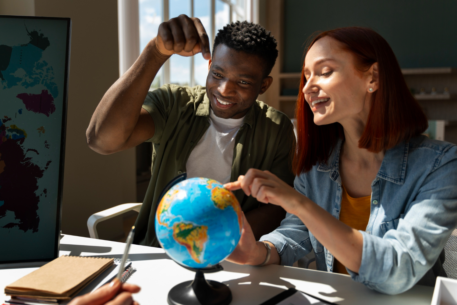 Foto de um homem negro e uma mulher branca sentados em uma mesa olhando e apontando para um globo, representando o curso Carreira de UX na Gringa.