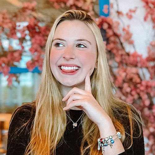 Foto de Karina Tronkos, apresentadora da DEXCONF 2024. Uma mulher branca, loira de cabelos lisos e compridos, sorrindo com a língua para fora e com o dedo indicador na bochecha.