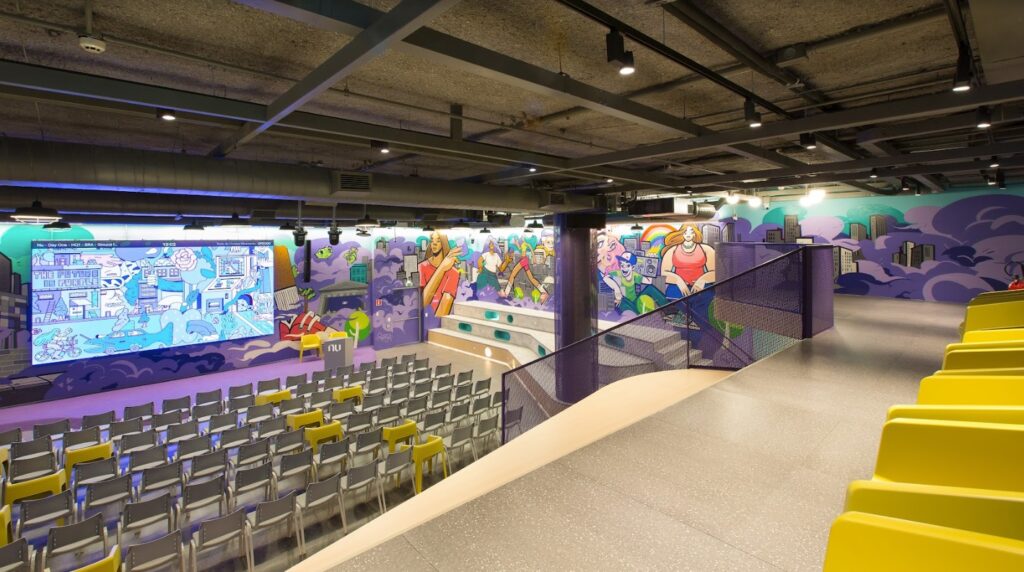 Foto do auditório do Nubank com palco, telão e cadeiras para participantes.